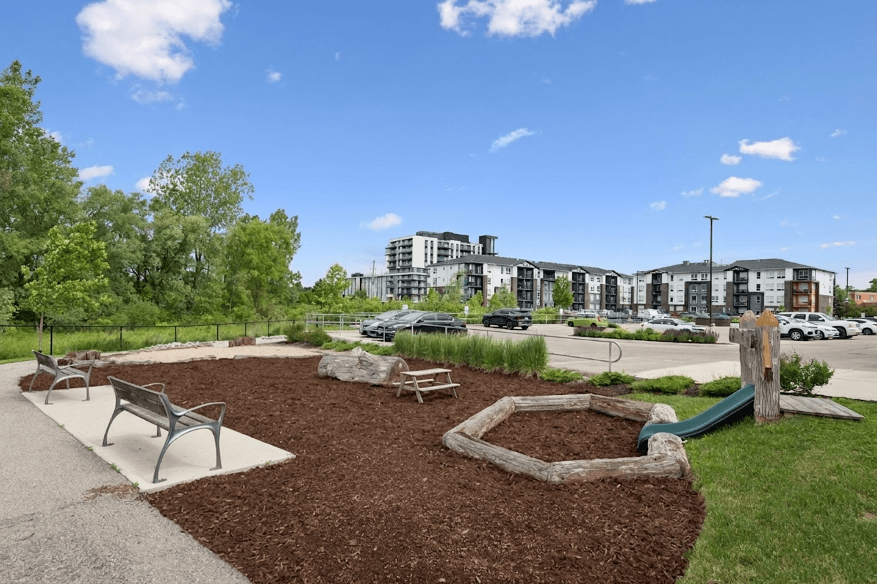 A park with a playground and benches in the foreground and apartment buildings in the background.
